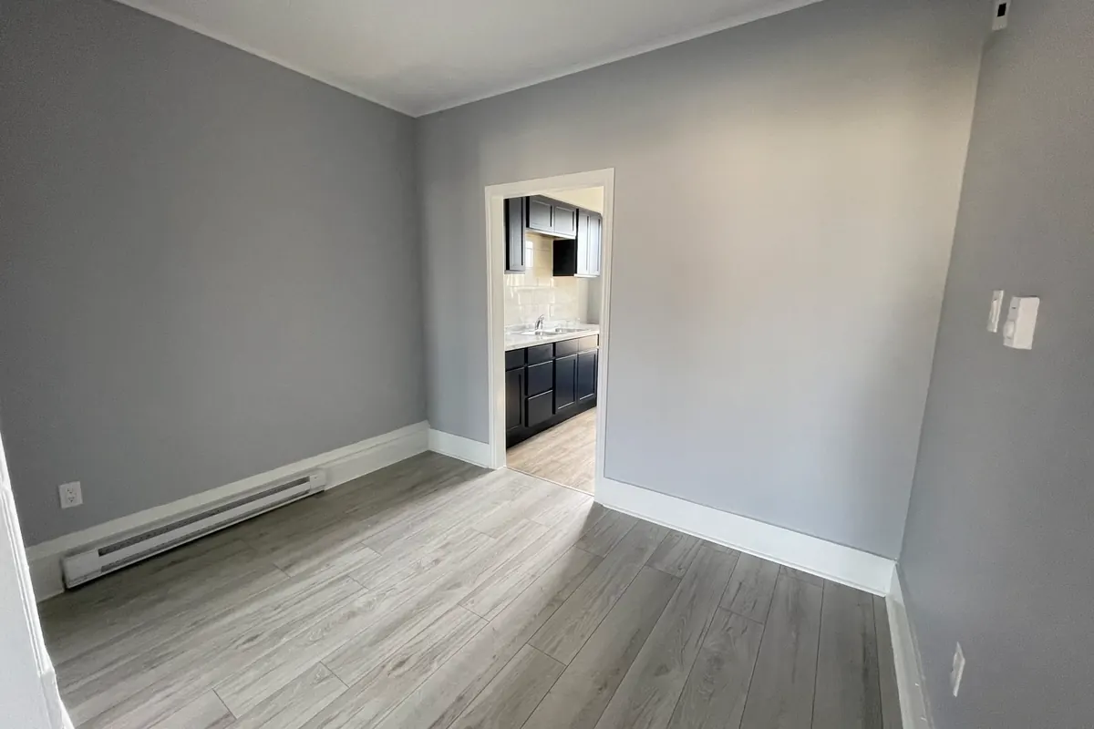 An empty grey room with plank floors and a doorway through to a navy kitchen.