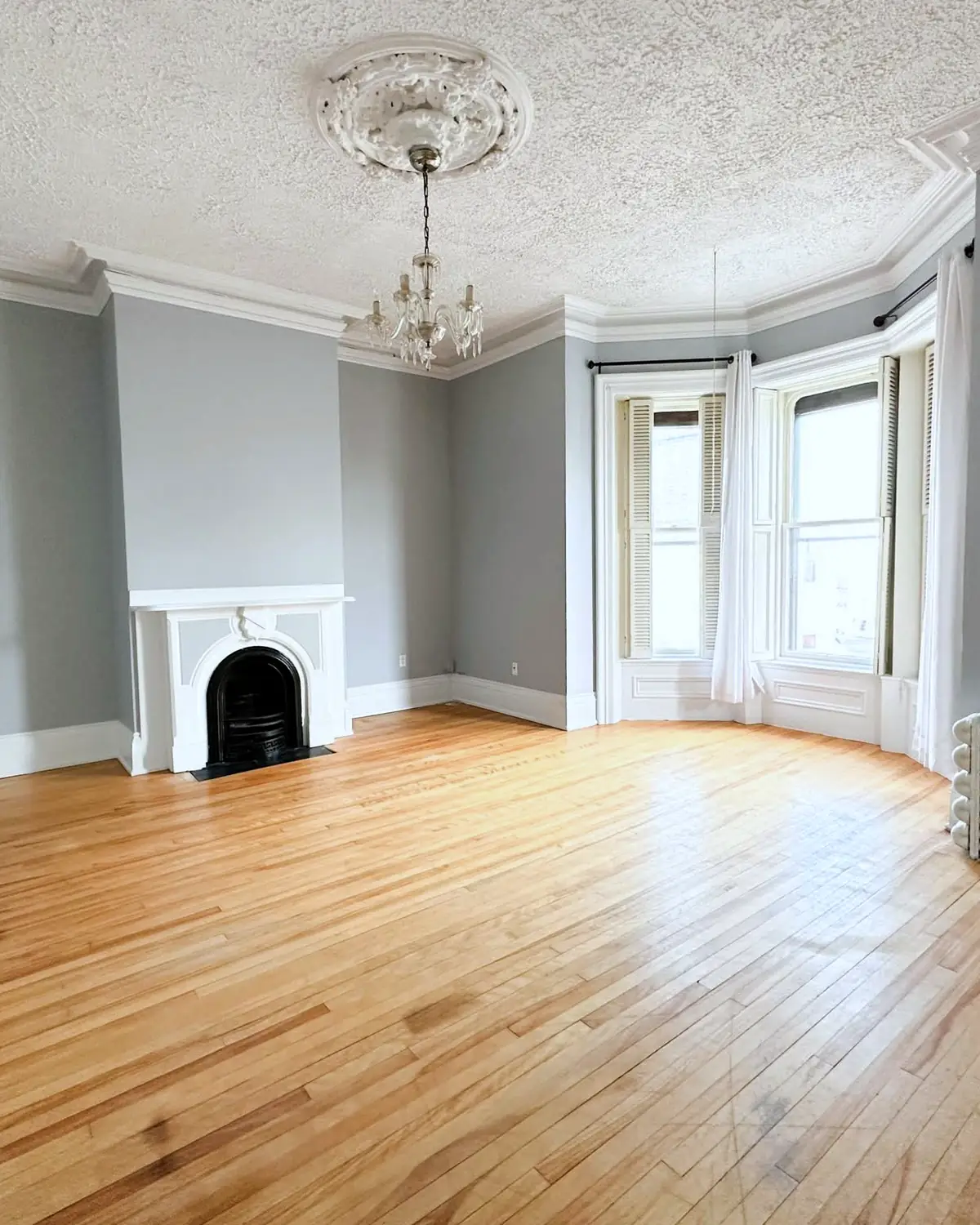 A renovated living room: hardwood floors, a white marble fireplace, a plaster ceiling medallion with a chandelier, and a bay window.